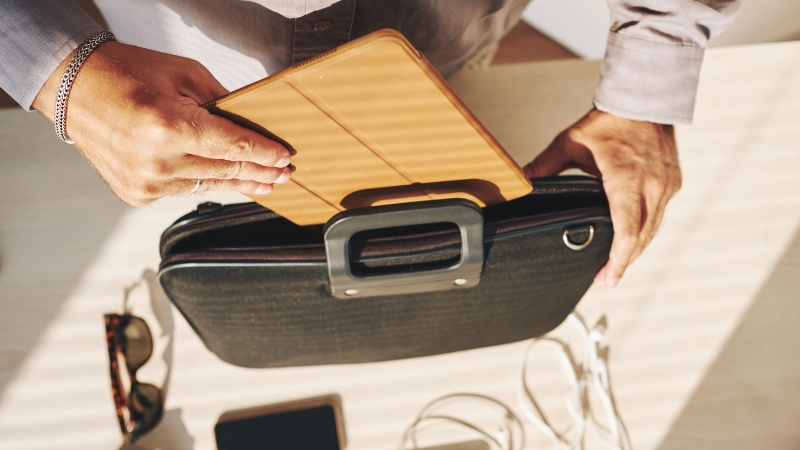 Muškarac ubacuje tablet u crnu poslovnu torbu na radnom stolu, pored naočara i telefona